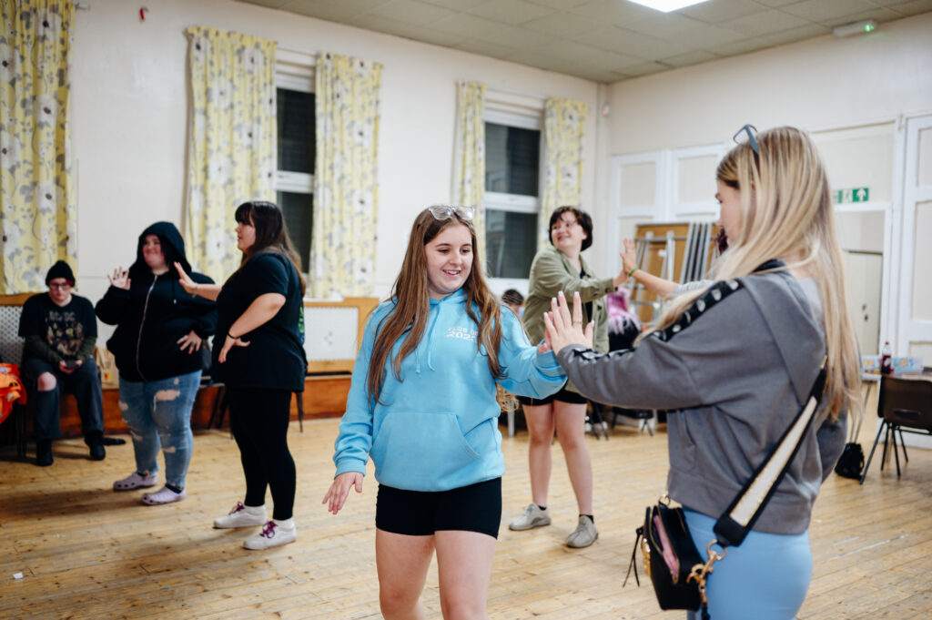 Ffotograff o grŵp o bobl ifanc, gyda merch ifanc mewn hwdi glas yn gwenu ac yn chwarae gyda ffrind, merch benfelen gyda hwdi llwyd arni mewn ystafell fawr gyda llenni melyn a gwyn.